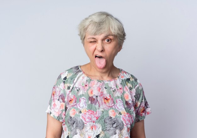 Joyful elderly woman blinks eye and sticks out tongue isolated on white wall