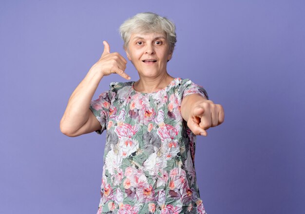 Joyful elderly woman gestures call me hand sign and points isolated on purple wall