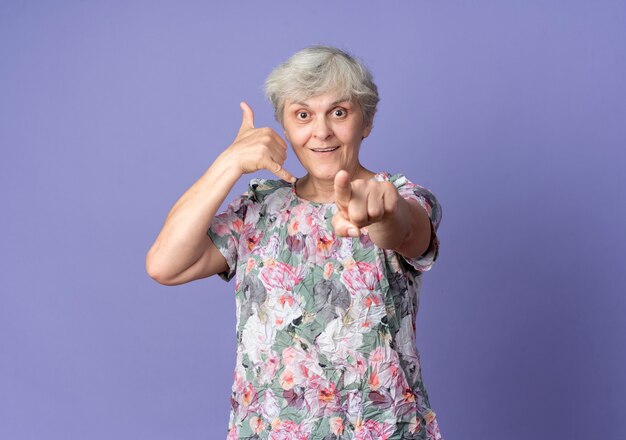 Joyful elderly woman gestures call me sign and points isolated on purple wall