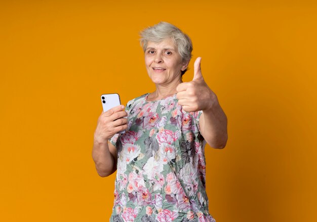 Joyful elderly woman holds phone and thumbs up isolated on orange wall