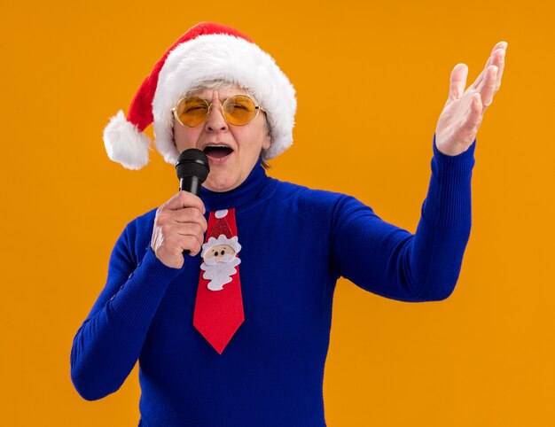 Joyful elderly woman in sun glasses with santa hat and santa tie holds mic pretending to sing isolated on orange background with copy space