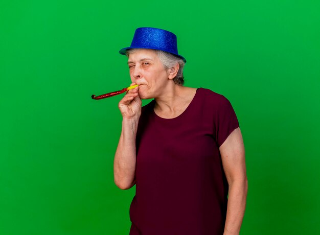 Радостная пожилая женщина в шляпе для вечеринки моргает, дует свисток на зеленый