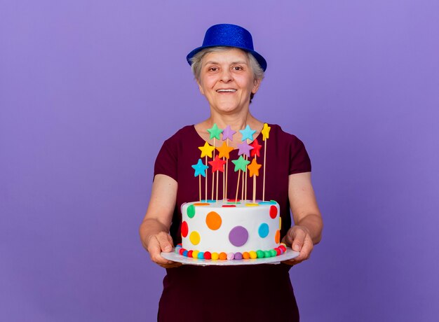 Joyful elderly woman wearing party hat holds birthday cake isolated on purple wall