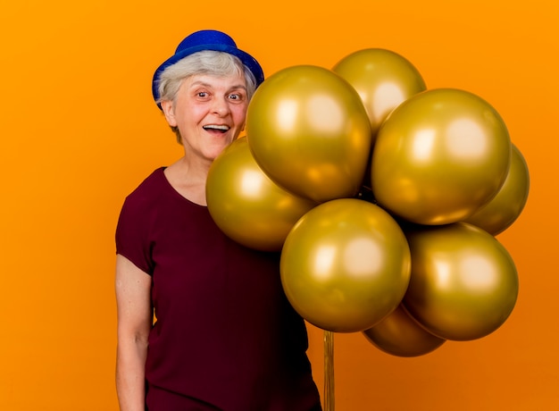 파티 모자를 쓰고 즐거운 노인 여성 복사 공간 오렌지 벽에 고립 된 헬륨 풍선을 보유