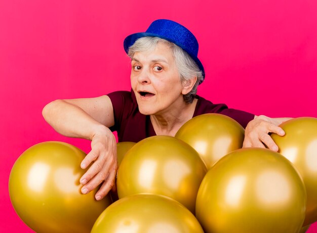 Радостная пожилая женщина в шляпе для вечеринки стоит за гелиевыми шарами на розовом