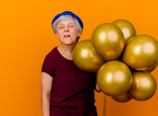 Joyful elderly woman wearing party hat stucks out tongue and holds helium balloons isolated on orange wall with copy space