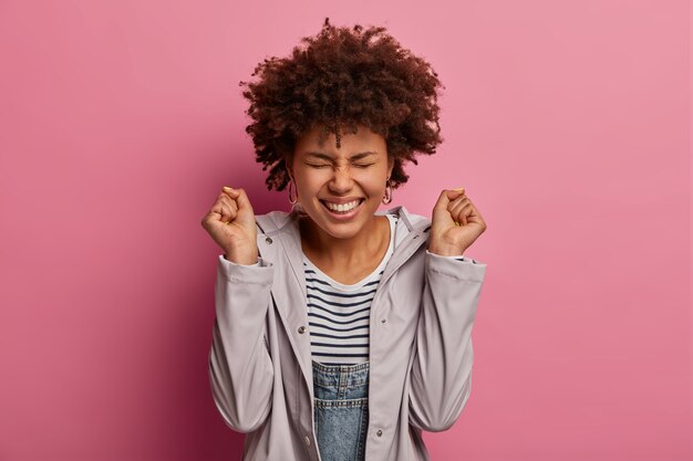 Joyful emotive ethnic curly woman clenches fists, celebates victory, smiles broadly, being in high spirit, closes eyes, wears casual anorak, rejoices achieve goal, poses against pink wall.