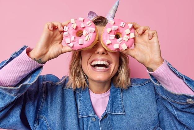 Joyful faceless woman covers eyes with big appetizing dougnuts with marshmallow smiles broadly enjoys eating sweet food wears stylish denim jacket isolated over pink background Delicious dessert