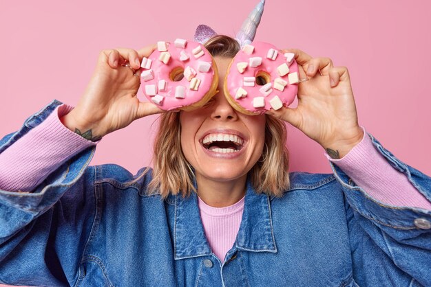 얼굴이 없는 유쾌한 여성이 마시멜로 미소로 식욕을 돋우는 커다란 도넛으로 눈을 가리고 분홍색 배경에 격리된 세련된 데님 재킷을 입고 달콤한 음식을 즐겨 먹는다. 맛있는 디저트