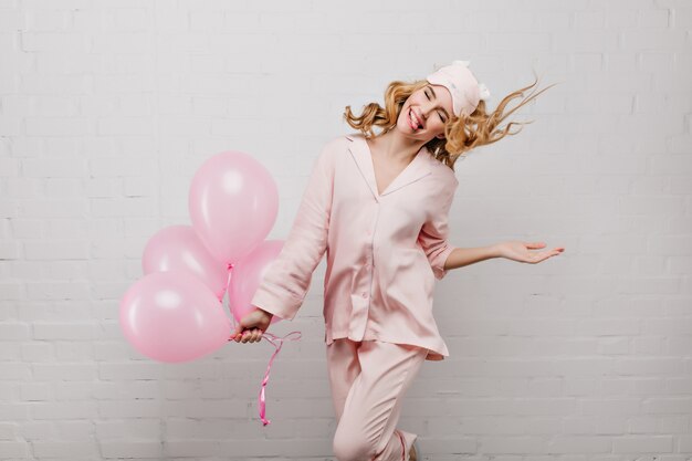 Joyful fair-haired girl posing with tongue out and holding bunch of pink balloons. Indoor portrait of ecstatic curly lady in pajamas and sleep mask celebrating birthday.