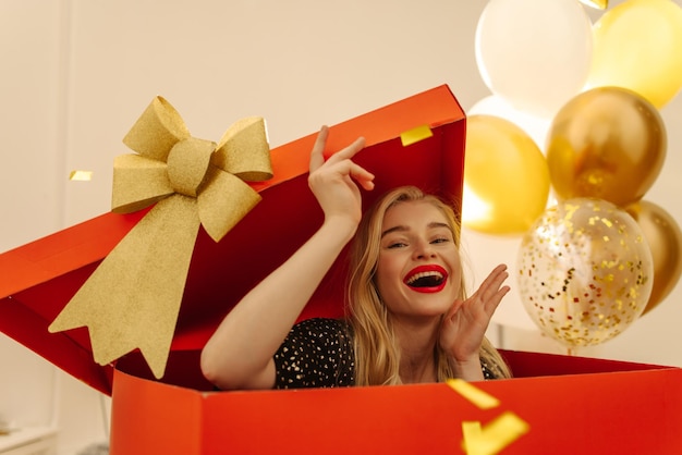 Радостная светлокожая молодая блондинка с энтузиазмом вылезает из большой красной коробки в качестве неожиданной концепции праздника