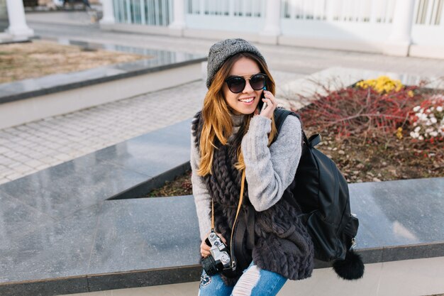 Joyful fashionable young woman in warm winter clothes, knitted hat, sunglasses sitting on street in city, speaking on phone. Travelling with backpack, camera, cheerful mood, positive emotions.