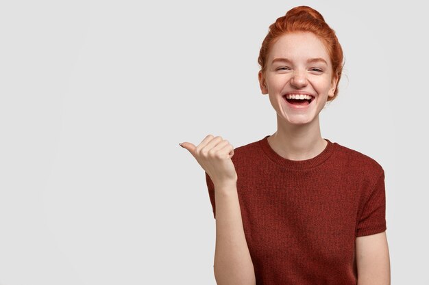 Joyful freckled red head youngster laughs positively, points aside with thumb,