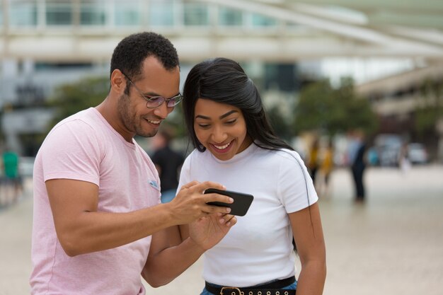 Joyful friends standing on street and using smartphone