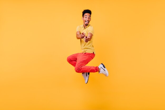 Joyful funny guy in white sneakers jumping. Indoor photo of laughing active african man.