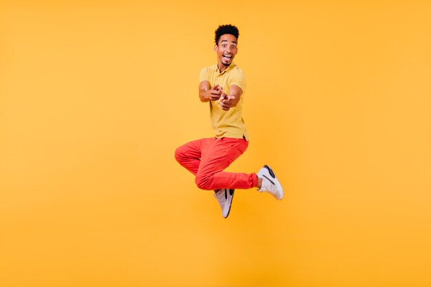 Радостный смешной парень в белых кроссовках прыгает. Фотография в помещении смеющегося активного африканского человека.