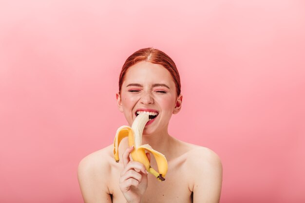 바나나를 먹는 즐거운 생강 소녀. 분홍색 배경에 열 대 과일을 즐기고 기쁘게 생각 된 여자의 전면 모습.