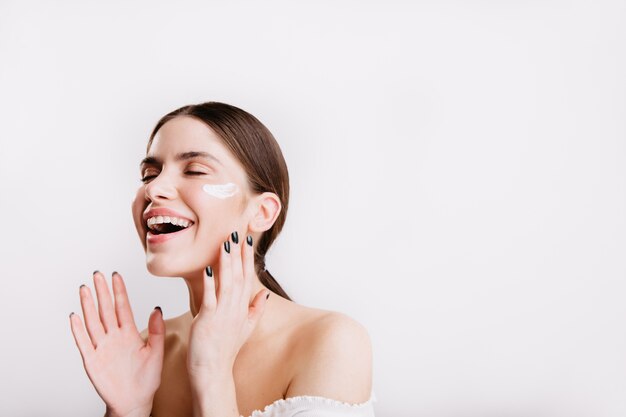 Joyful girl enjoys applying moisturizing cream to clean face. Brunette without makeup laughs on white wall.