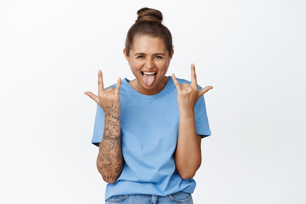 Joyful girl having fun, showing rock on heavy metal horns, sticking tongue and shouting excited, standing in blue t-shirt on white