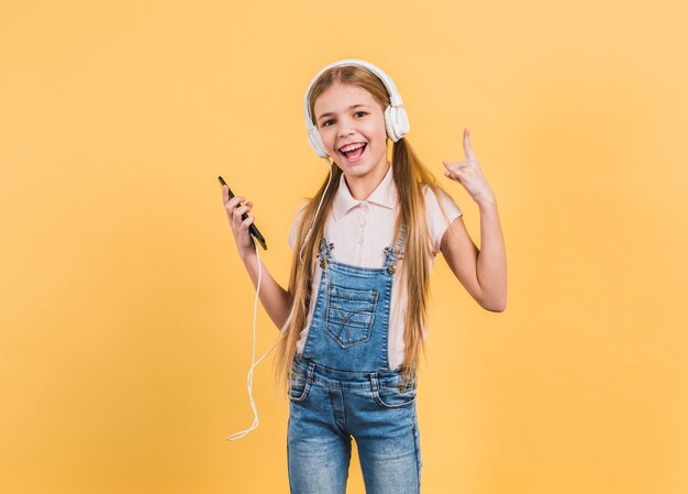 Joyful girl listening music on headphone making rock sign against yellow background