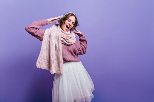 Joyful girl in lush white skirt looking down with inspired smile. refined brunette lady in long warm scarf dreamy posing on purple wall.