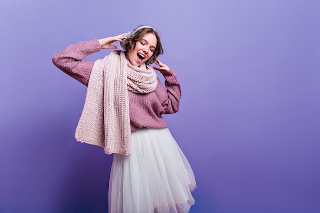 Joyful girl in lush white skirt looking down with inspired smile. refined brunette lady in long warm scarf dreamy posing on purple wall.