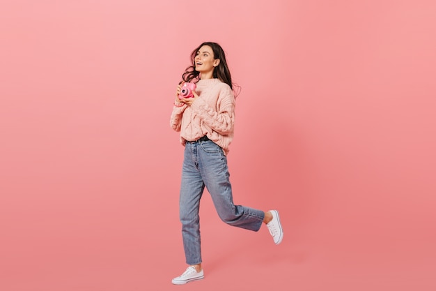 Joyful girl in moms jeans runs on pink background. Dark haired girl posing with instax camera.