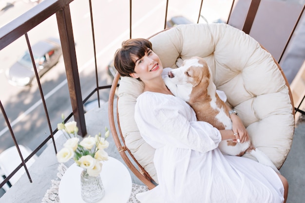 Free Photo joyful girl sitting in chair beside vase of white roses and stroking beagle dog. beautiful brown-haired woman enjoying fresh air on balcony with pet lies on her knees