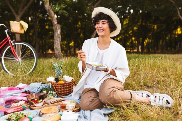 Joyful girl sitting in hat and white shirt with salad in hands happily laughing spending time on picnic in park