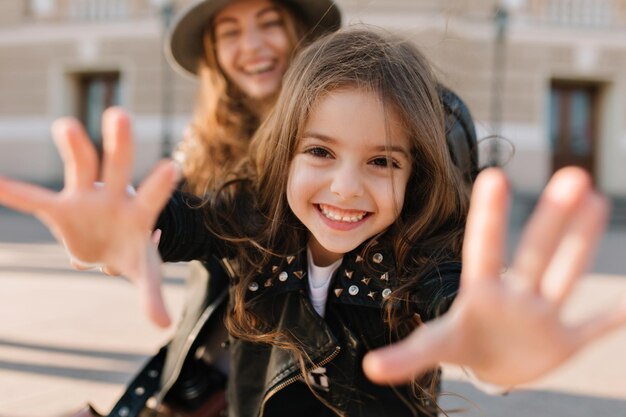 Joyful girl smiles charmingly and extends her hands to the camera, while her mother standing behind. Happy beautiful woman in hat spending time with excited daughter and having fun outside on weekend.