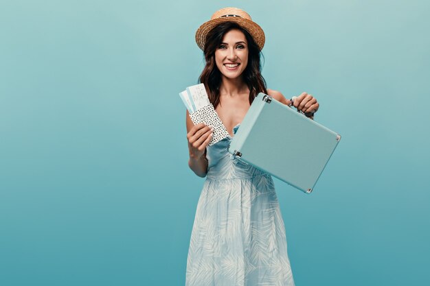 Joyful girl in straw hat holding tickets and suitcase on blue background