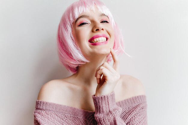 Joyful girl with trendy bright makeup laughing on white wall. Close-up photo of blissful young woman in pink peruke smiling while posing in purple sweater.