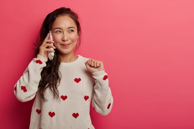 Joyful girlfriend enjoys casual friendly conversation with pal, talks and rejoices cool news, looks aside with dreamy expression, raises clenched fist, keeps smartphone pressed to ear.