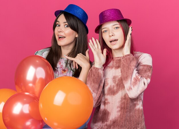 Joyful  girls wearing party hat holding balloons showing phone call gesture isolated on pink wall