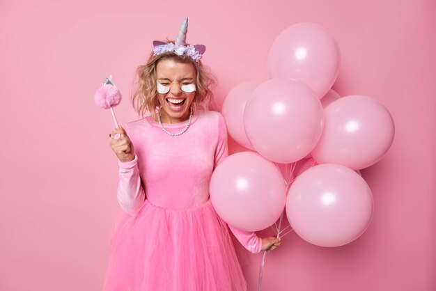 Joyful glad woman wears dress unicorn headband applies beauty patches holds bunch of inflated balloons has fun on party celebrates special occasion poses against pink background Celebration