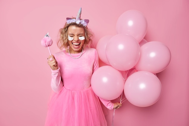 Радостная радостная женщина носит платье, повязка на голову единорога, накладывает косметические пластыри, держит кучу надутых воздушных шаров, веселится на вечеринке, празднует особые позы на розовом фоне.