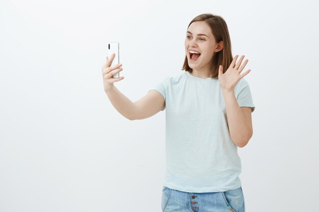 joyful good-looking woman talking via smartphone video message waving palm, smiling broadly at device screen saying hello communicating via internet