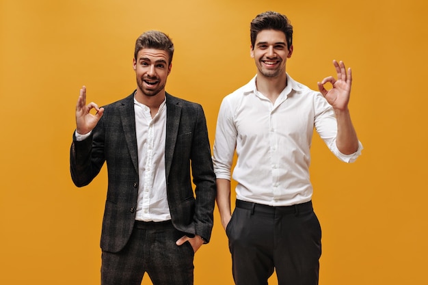 Free photo joyful goodhumored businessmen in white shirts and black pants smile sincerely and show ok signs on orange isolated background