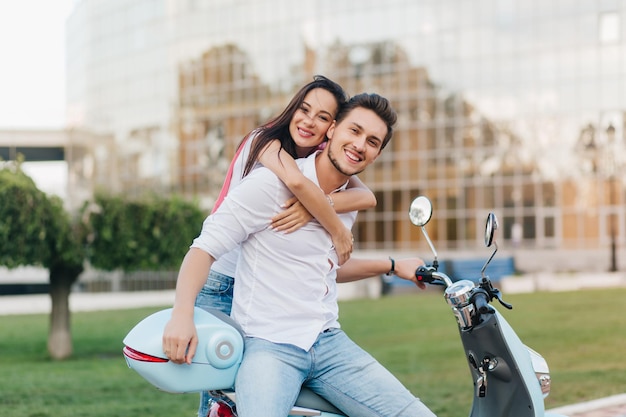 Joyful guy in jeans and shirt posing while his attractive girlfriend huggs him from behind. Outdoor portrait of smiling romantic couple standing in front of modern building after riding on scooter.