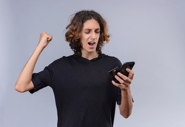 Free photo joyful guy with long hair in black t-shirt holding a phone and a card on white wall