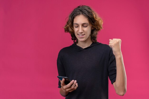 Радостный парень с длинными волосами в черной футболке держит телефон на розовой стене