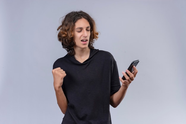 Joyful guy with long hair in black t-shirt holding a phone on white wall