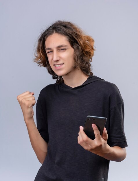 Радостный парень с длинными волосами в черной футболке держит телефон на белой стене