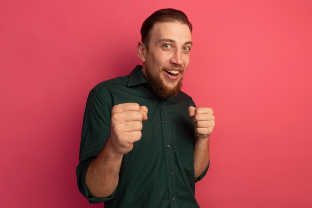 Joyful handsome blonde man keeps fists isolated on pink wall