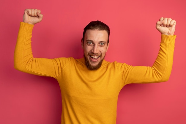 Joyful handsome blonde man raising fists up on pink