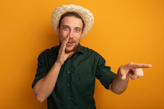 Joyful handsome blonde man with beach hat holds hand close to mouth and points at side isolated on orange wall