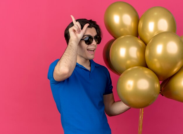 Joyful handsome caucasian man in sun glasses holds helium balloons and gestures loser sign 