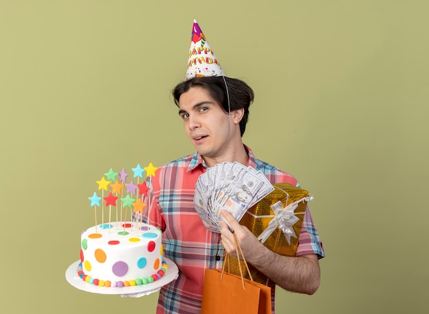 Un bell'uomo caucasico allegro che indossa un berretto da compleanno tiene in mano una scatola regalo in carta per la spesa, soldi e una torta di compleanno