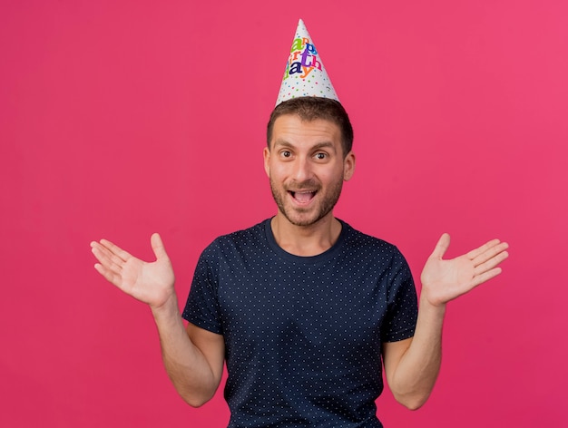 Joyful handsome caucasian man wearing birthday cap stands with raised hands isolated on pink background with copy space