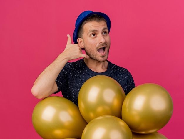 Joyful handsome caucasian man wearing blue party hat gestures call me sign and holds helium balloons looking at side isolated on pink background with copy space